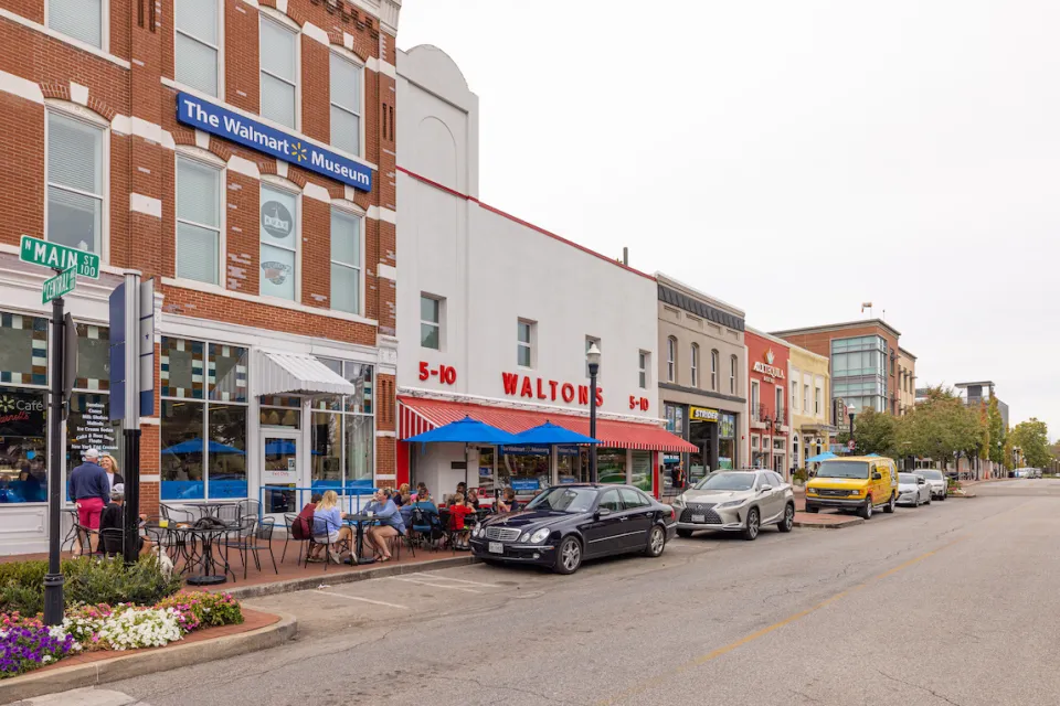 Main Street in Bentonville, Arkansas with three-story brick buildings, people sitting at tables on sidewalk, and sign on building for "The Walmart Museum."